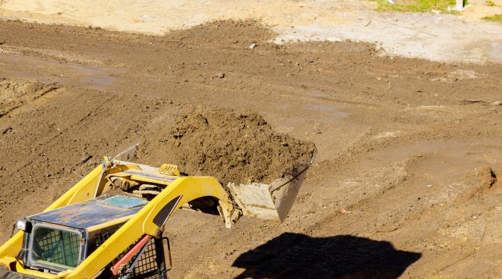 A yellow skid steer loader using a grading bucket to move and level soil on a construction site, casting a sharp shadow in the bright sun.