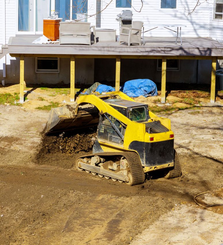 A yellow compact track loader moving soil in a backyard next to a house with a wooden deck, preparing the area for landscaping or drainage.