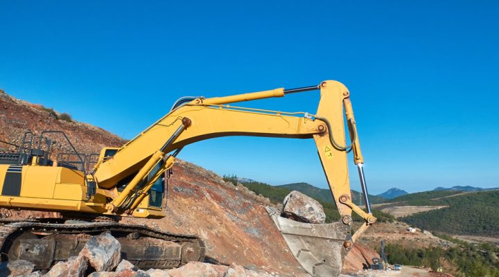 Large yellow excavator lifting a massive boulder at a rocky quarry site under a clear blue sky.