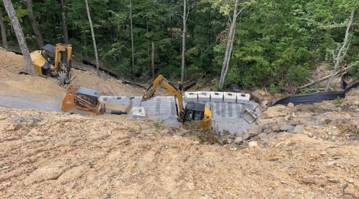 Three pieces of heavy equipment working simultaneously on a steep, wooded slope to install concrete foundation blocks for a new structure.