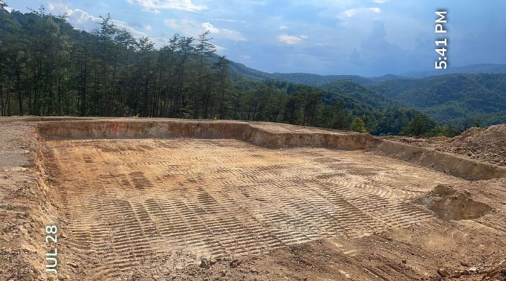 A vast, meticulously graded building pad on a mountaintop with tire tracks across the soil, offering panoramic views of forested hills under a blue sky at 5:41 PM.