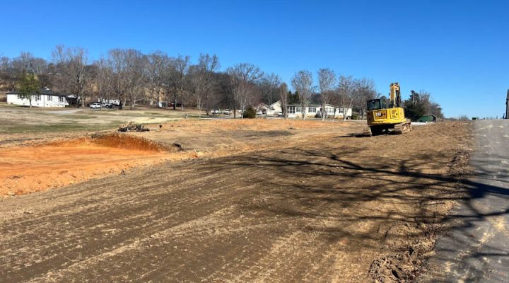 A yellow excavator parked on a graded roadside area next to a paved road, with a backdrop of suburban homes and bare trees under a clear blue sky.