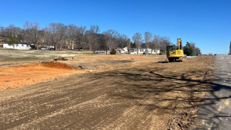 A yellow excavator parked on a graded roadside area next to a paved road, with a backdrop of suburban homes and bare trees under a clear blue sky.