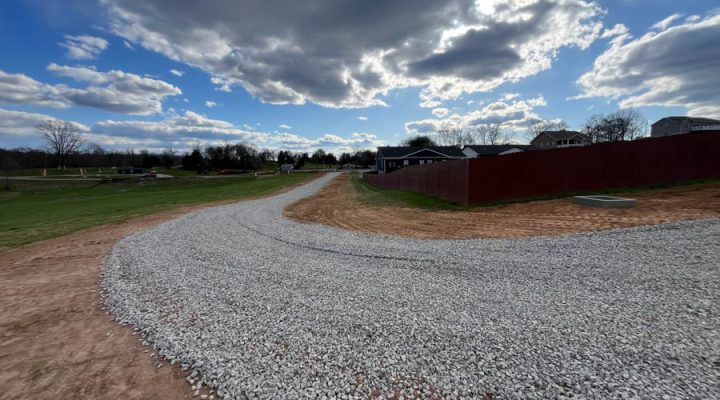 A winding gravel driveway bordered by freshly graded earth, leading towards a residential area under a dramatic sky with scattered clouds.