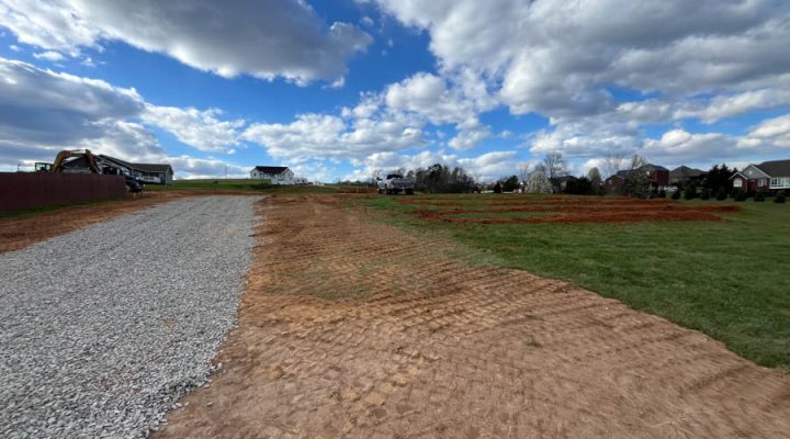 A newly installed gravel driveway running alongside a freshly graded, tire-tracked dirt area, with construction equipment and houses visible in the distance under a dramatic cloudy sky.