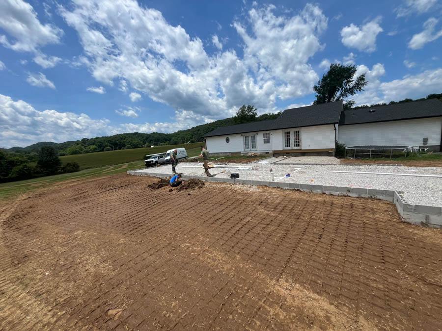 Two workers installing utilities on a large, graded lot with a white house in the background, under a vibrant blue sky with fluffy white clouds.