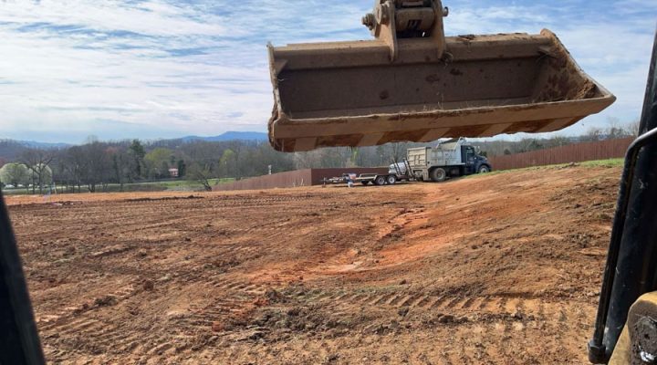 View from inside an excavator looking at its bucket raised high, with a dump truck parked on a large, graded red clay field ready to receive soil.