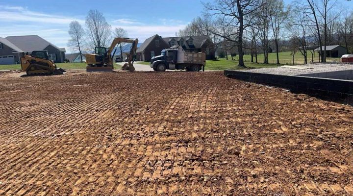 A freshly graded residential lot with visible tire tracks, featuring a skid steer, excavator, and dump truck near new homes under construction on a sunny day.