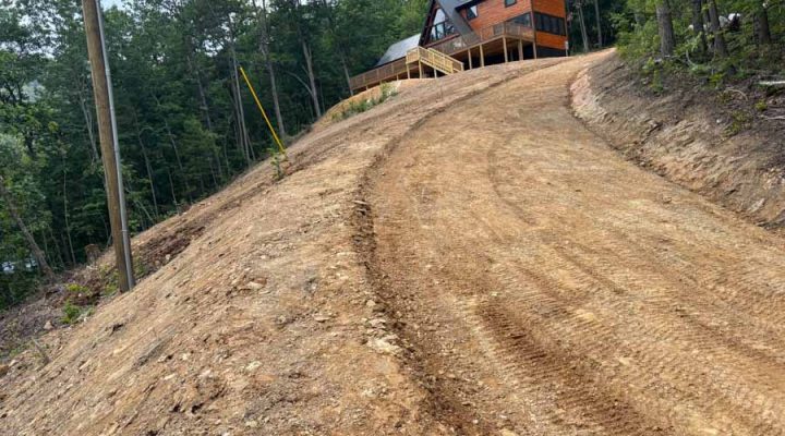 A freshly graded dirt driveway winding up a steep hill to a modern wooden cabin, surrounded by dense forest under a cloudy sky.