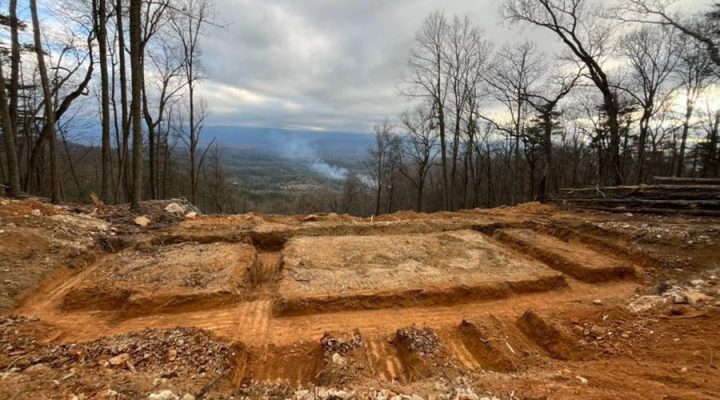 Deep, red clay foundation trenches carved into a mountaintop site, surrounded by bare winter trees and offering a sweeping view of a distant valley under an overcast sky.