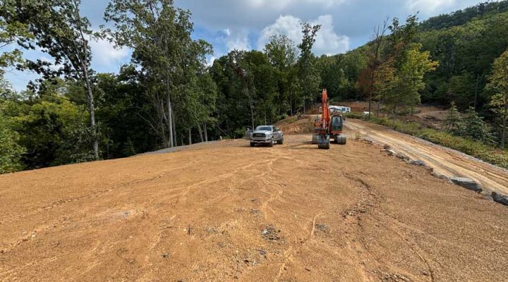 A wide, graded lot with tire tracks, featuring an orange excavator and a pickup truck parked nearby, surrounded by dense trees and distant mountains under a cloudy sky.
