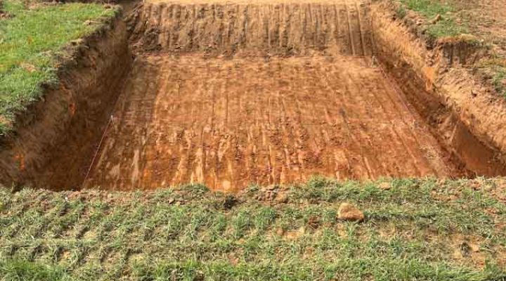 A deep, rectangular excavation for a home’s foundation, with clean, straight walls and tire tracks on the compacted soil, located in front of a modern brick house.
