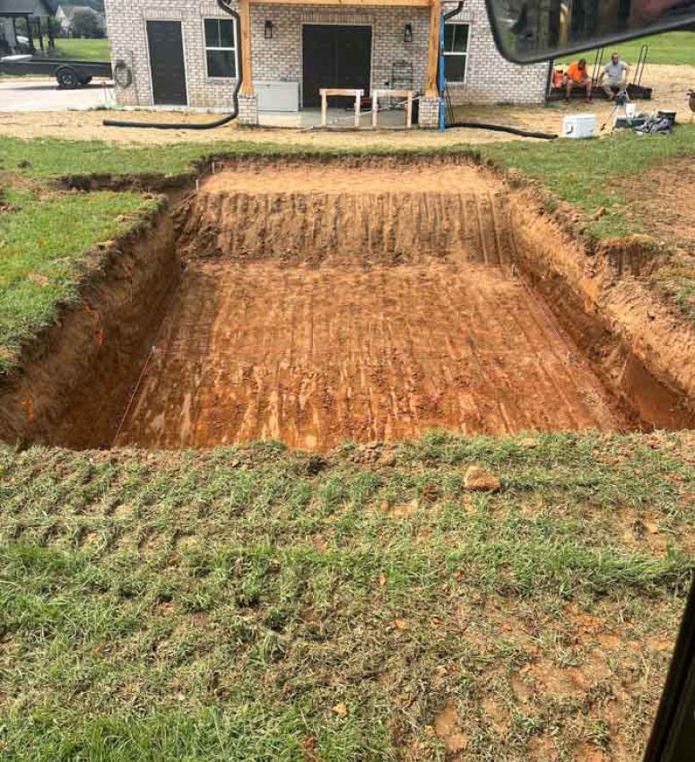 A deep, rectangular excavation for a home’s foundation, with clean, straight walls and tire tracks on the compacted soil, located in front of a modern brick house.