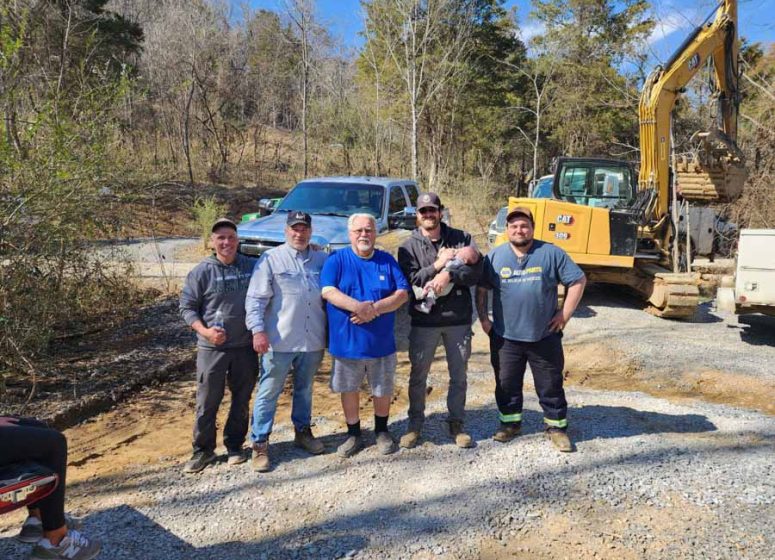 Five members of the Rooney Excavating crew posing for a photo on a gravel site, with an excavator and truck in the background under a sunny blue sky.