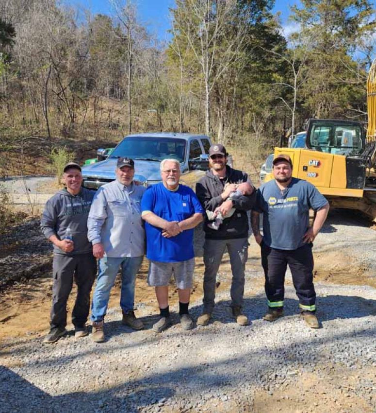 Five members of the Rooney Excavating crew posing for a photo on a gravel site, with an excavator and truck in the background under a sunny blue sky.