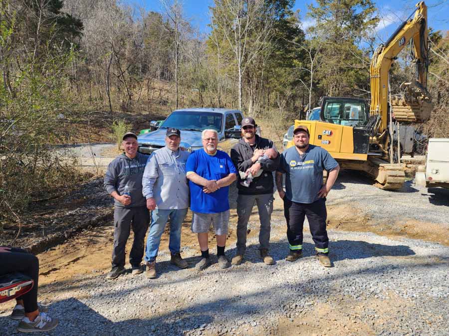 Five members of the Rooney Excavating crew posing for a photo on a gravel site, with an excavator and truck in the background under a sunny blue sky.
