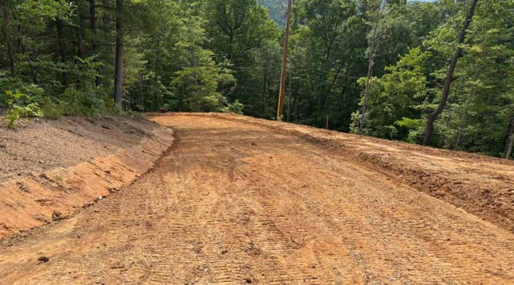 A winding, freshly graded dirt road cutting through a dense forest, showing clear tire tracks and a view of distant mountains under a partly cloudy sky.