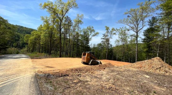 An orange skid steer loader working on a cleared, flat building pad at the edge of a wooded area, with a pile of excavated earth nearby under a clear sky.