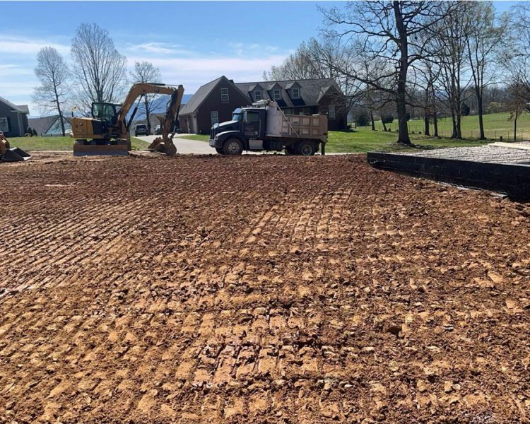 A freshly graded residential lot with visible tire tracks, featuring a skid steer and excavator on-site near new homes under construction.
