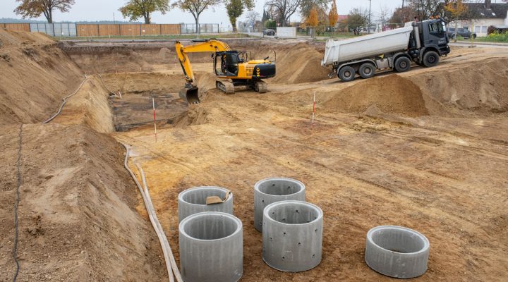 Wide shot of a construction site featuring an excavator, a dump truck, and precast concrete rings ready for installation.