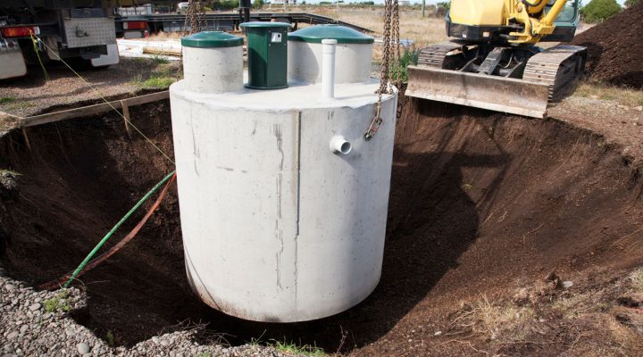 A large concrete septic tank being carefully lowered into a prepared excavation pit with an excavator nearby.